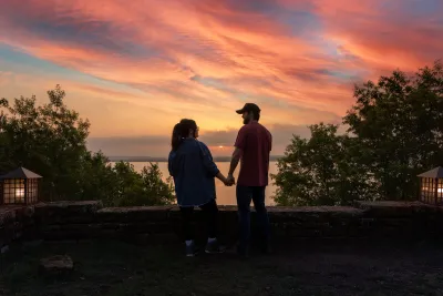 A couple holding hands at a stone overlook, watching a pink and orange sunset over Lake Brownwood