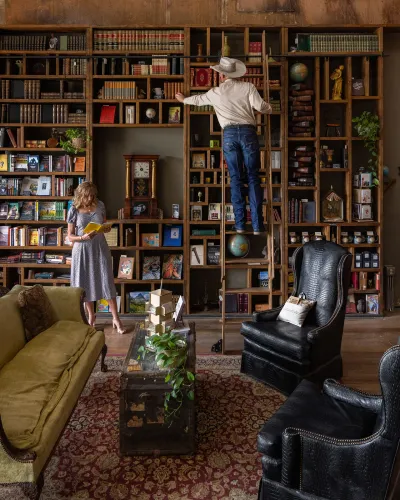 A woman browsing a book and a man in a cowboy hat on a rolling ladder inside a downtown Brownwood shop lined floor to ceiling with bookshelves