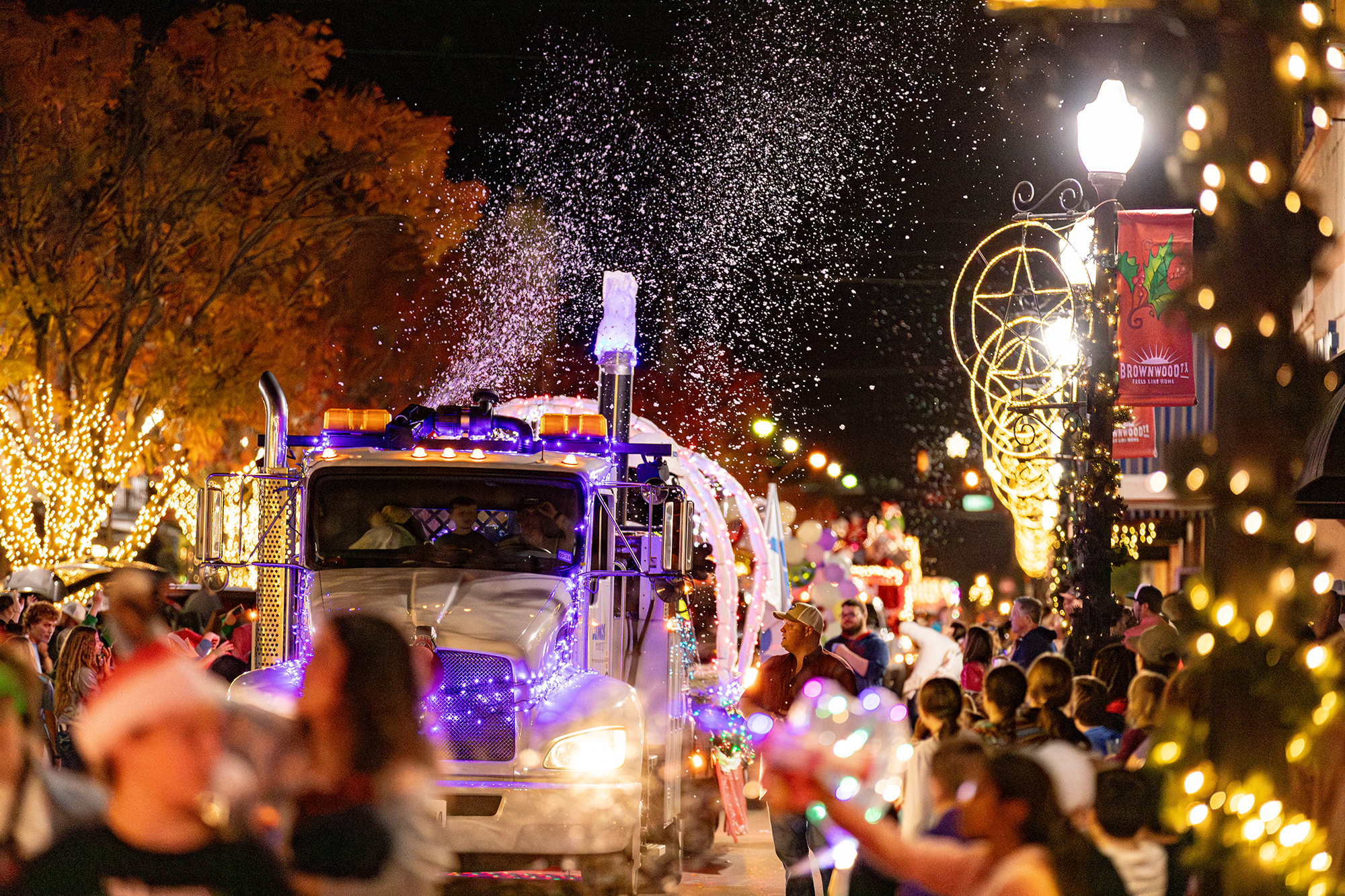 Photo of a Water Semi Truck decorated as a parade float with lights and blowing fake snow bubbles in front of the crowd lining the streets during the lighted christmas parade.