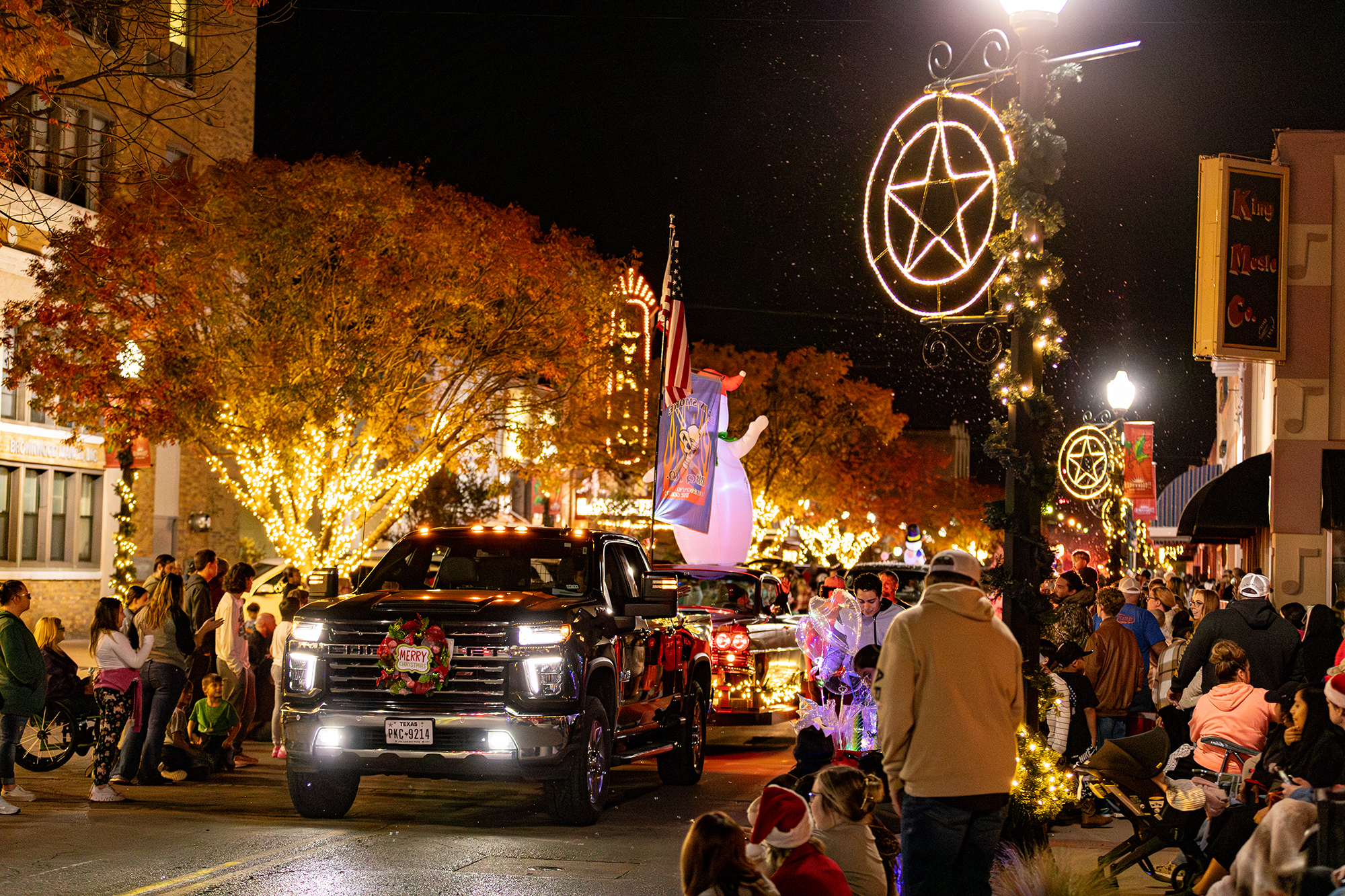 Photo of the lighted christmas parade with floats being pulled by trucks with the streets lined with a crowd looking at the lighted street and parade.