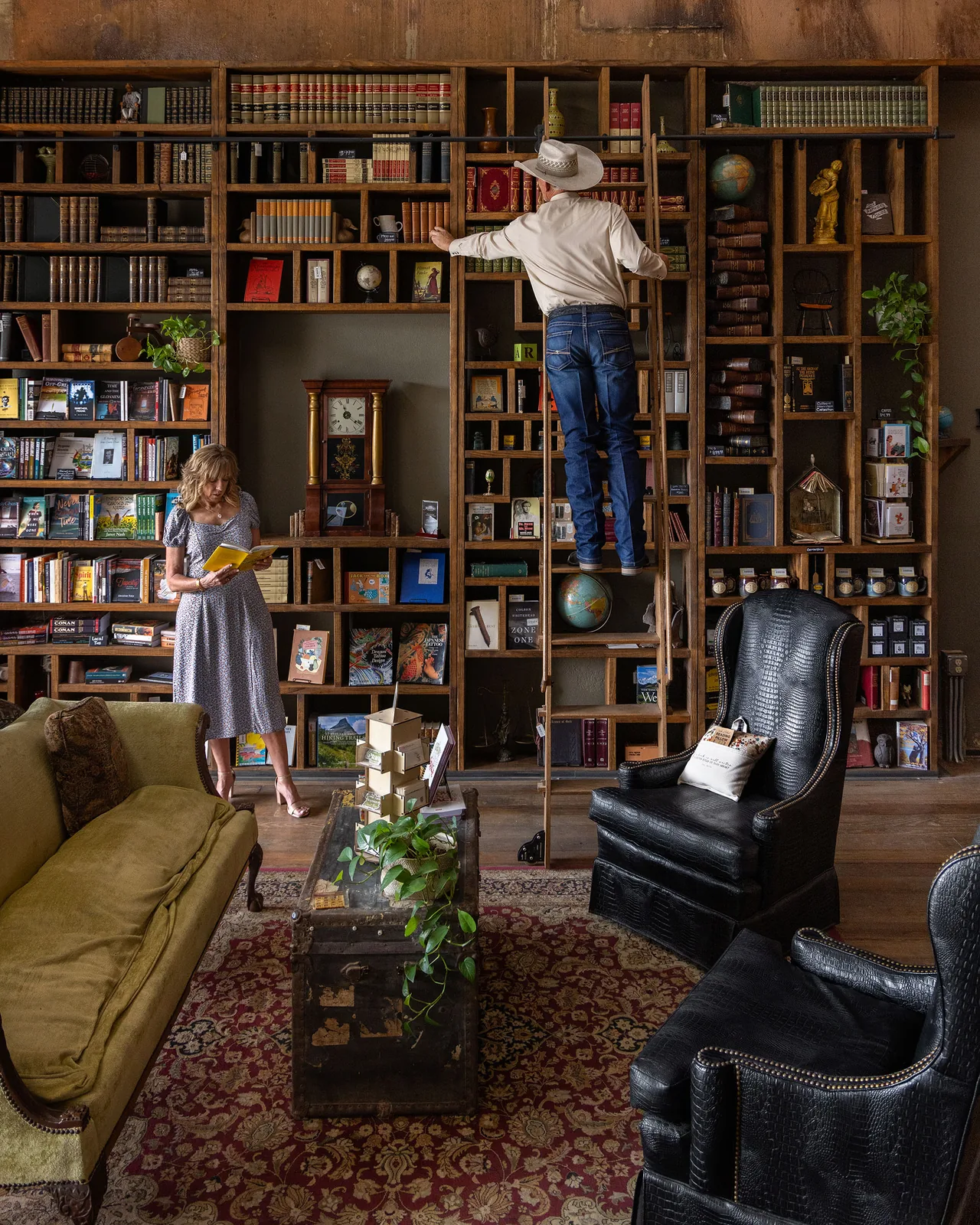 A woman browsing a book and a man in a cowboy hat on a rolling ladder inside a downtown Brownwood shop lined floor to ceiling with bookshelves