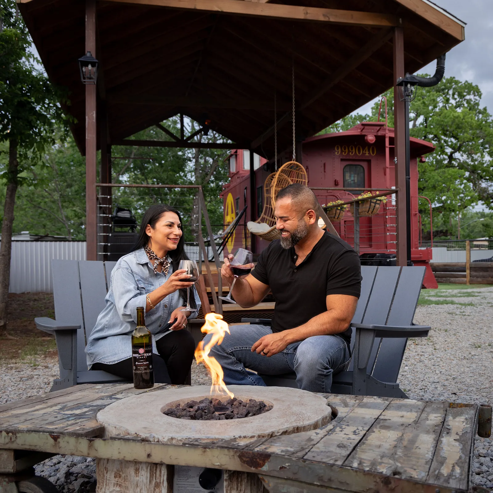 A couple toasting glasses of red wine beside a fire pit, with a red caboose parked behind them