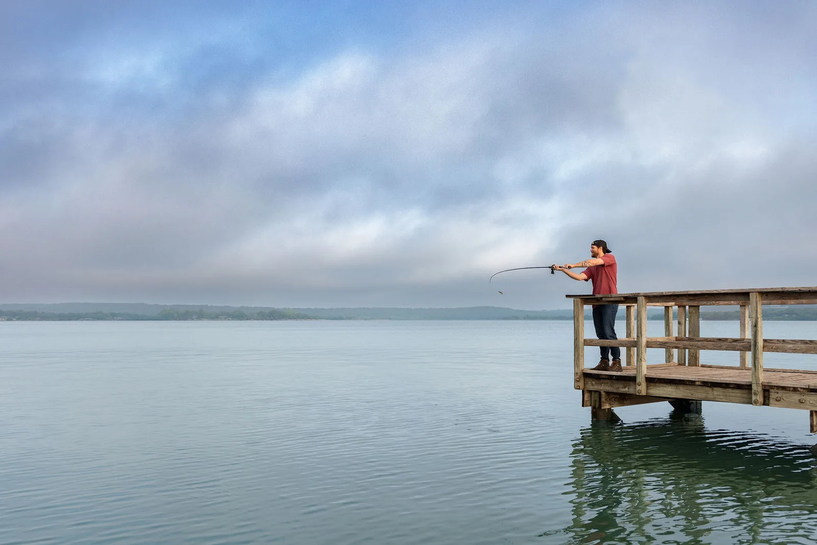 An angler casting from a wooden fishing pier on a still, misty Lake Brownwood